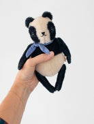 A small plush panda toy with black and cream fur and a light blue bow tie, held in a person's hand against a plain light gray background.